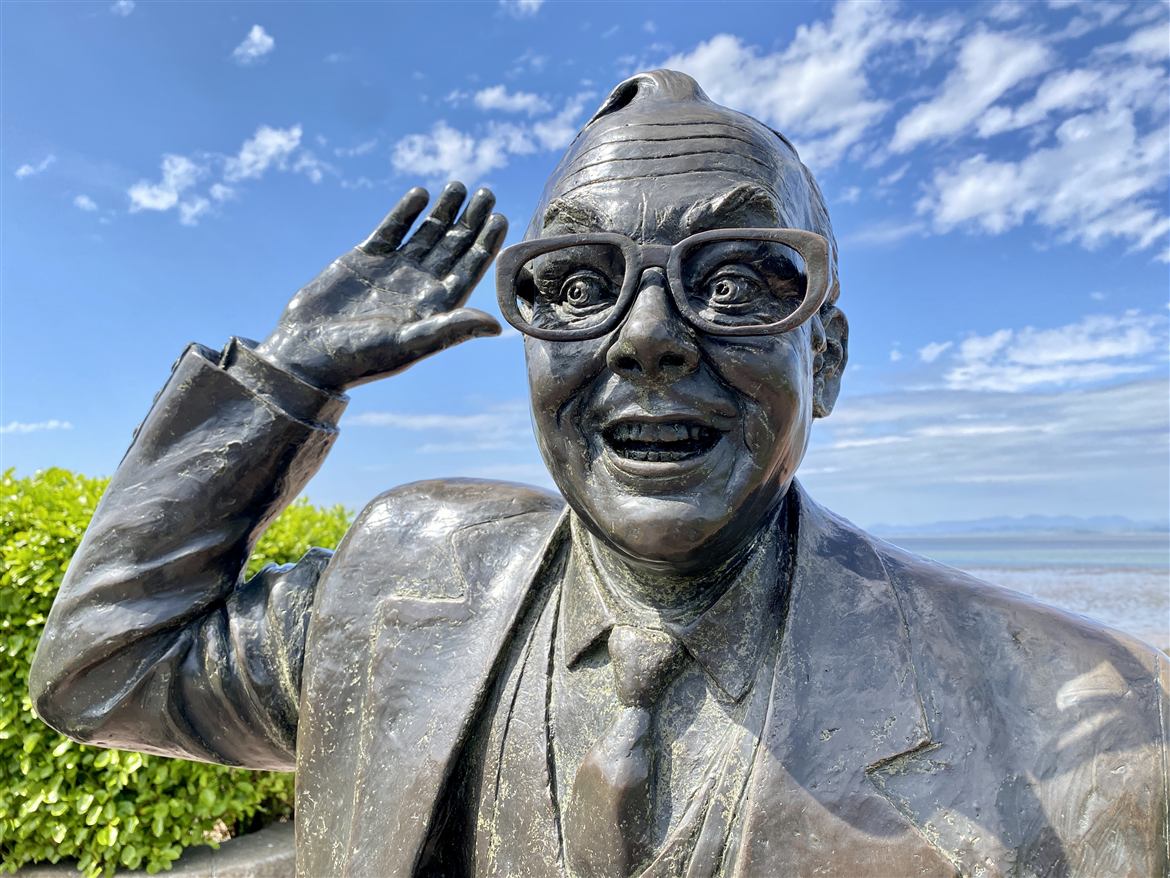 Eric Morecambe statue mid-dance on Morecambe promenade with Morecambe Bay and a blue sky behind