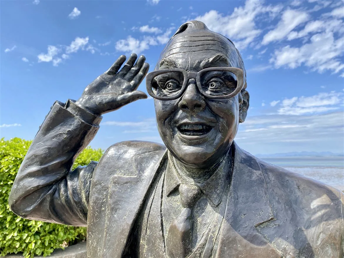 Eric Morecambe statue mid-dance on Morecambe promenade with Morecambe Bay and a blue sky behind